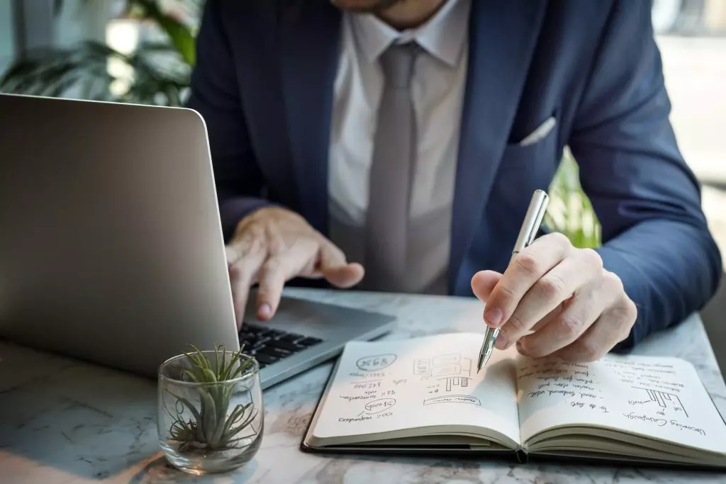 A man writing in a notebook while looking at a laptop