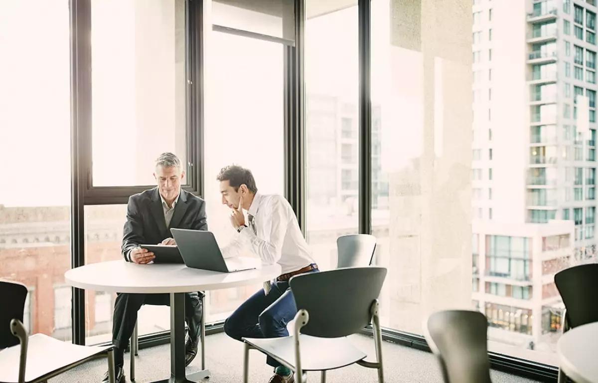 Real estate investors reviewing joint venture real estate agreement on digital tablet in office with large windows overlooking commercial real estate