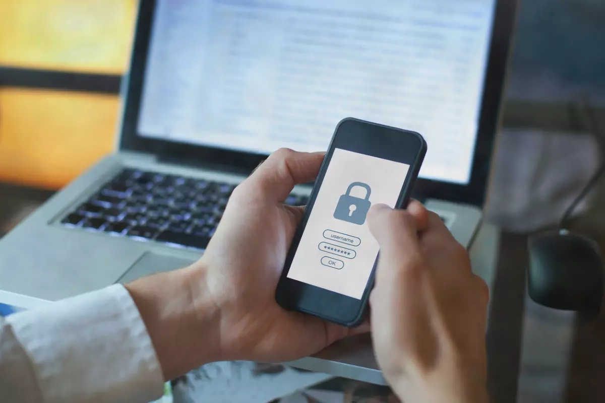 Hand holding a smartphone with a screen showing a locked icon with a laptop in the background.