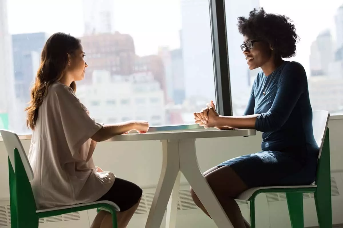 Two women talking at a table by a window.