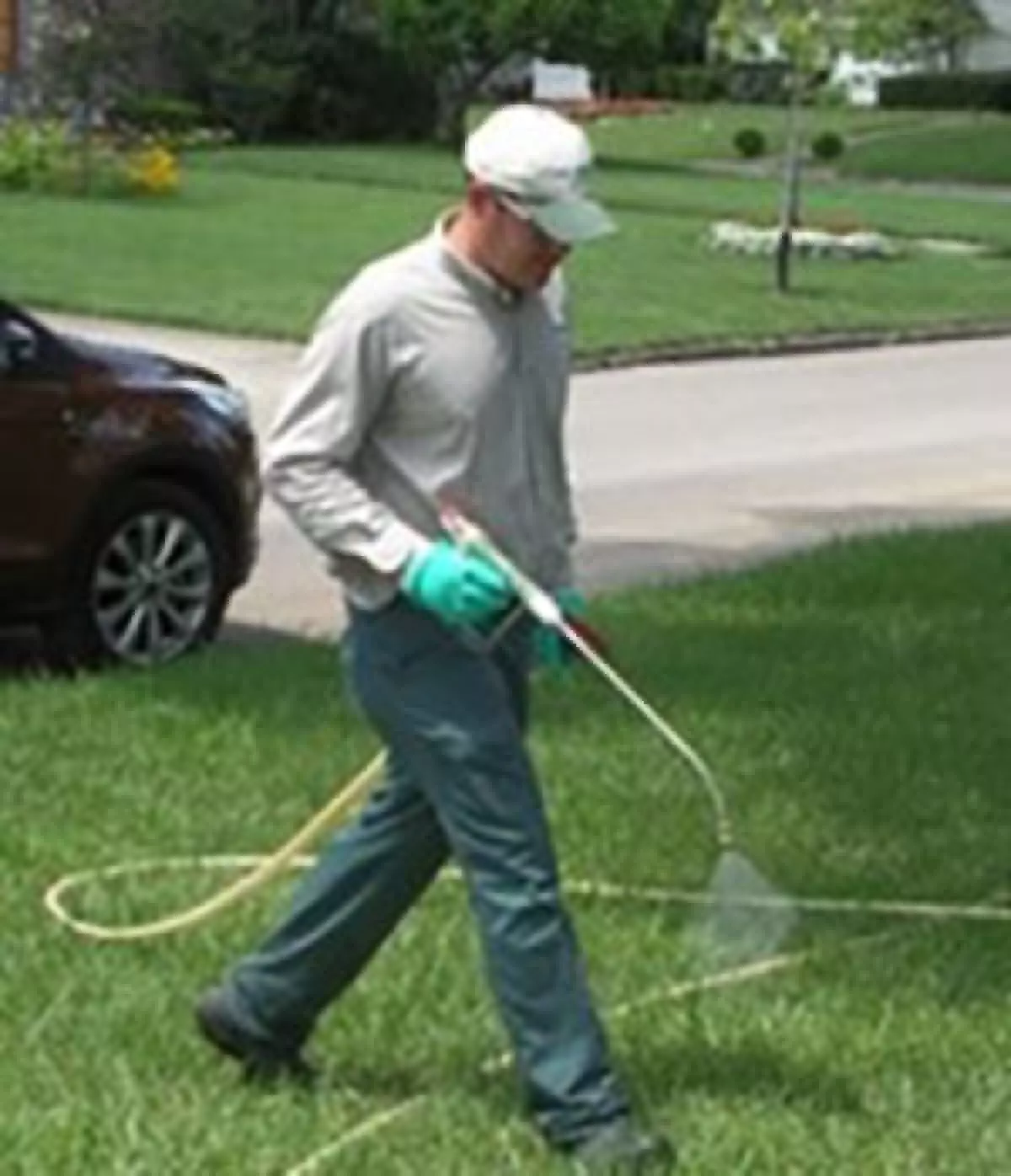 Man wearing gloves, long pants, long-sleeved shirt, and baseball cap spraying lawn with wand.